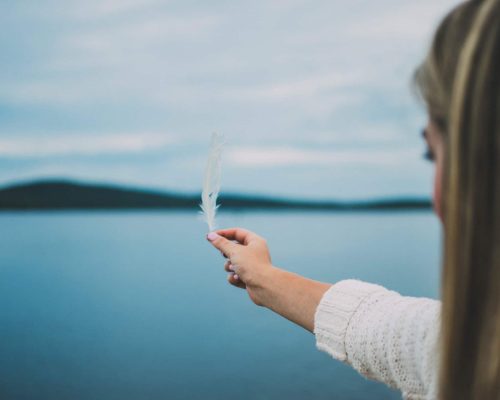 Imagem de uma pessoa segurando uma pena perto de um lago tranquilo.